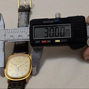 May include: A gold-tone wristwatch with a square face and a brown leather band. The watch face is white with black hands and black Roman numeral hour markers. The watch is being measured with a digital caliper, which reads 30.00 mm.