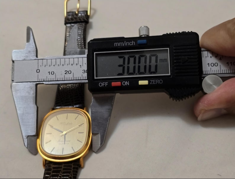 May include: A gold-tone wristwatch with a square face and a brown leather band. The watch face is white with black hands and black Roman numeral hour markers. The watch is being measured with a digital caliper, which reads 30.00 mm.