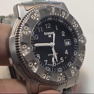Puede incluir: Un reloj Traser P6500 Type 3 plateado con una esfera negra y marcas blancas. El reloj tiene un bisel giratorio con n&uacute;meros del 0 al 60. La esfera del reloj tiene una ventana de fecha en la posici&oacute;n de las 3 en punto y las palabras "traser P65" impresas en ella. El reloj es resistente al agua hasta 200 metros.