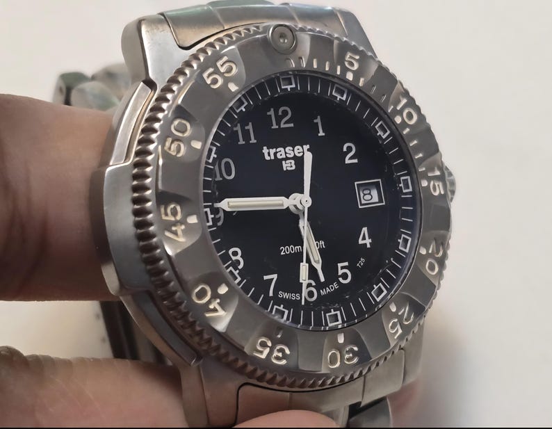 Puede incluir: Un reloj Traser P6500 Type 3 plateado con una esfera negra y marcas blancas. El reloj tiene un bisel giratorio con n&uacute;meros del 0 al 60. La esfera del reloj tiene una ventana de fecha en la posici&oacute;n de las 3 en punto y las palabras "traser P65" impresas en ella. El reloj es resistente al agua hasta 200 metros.