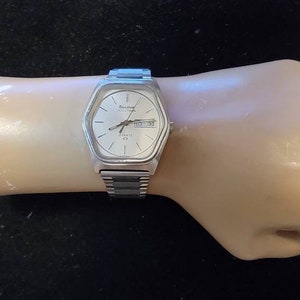 Op de afbeelding: Een zilverkleurig polshorloge met een witte wijzerplaat en een dag- en datumaanduiding. Het horloge heeft een zilveren metalen band.