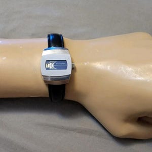 May include: A wristwatch featuring a silver-toned rectangular case with rounded edges and a dark blue dial face. The watch has a black leather strap and a small silver crown on the right side. The design is sleek with a minimalist display.