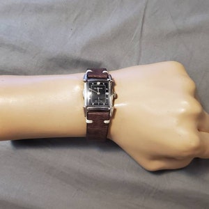 Op de afbeelding: Een zilveren horloge met een zwarte wijzerplaat en een bruin leren bandje. Het horloge zit om de pols van een persoon.