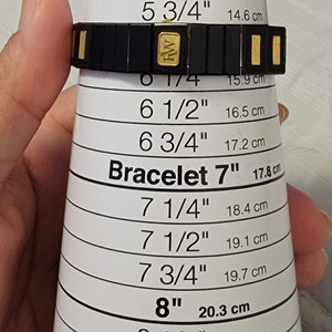 May include: A black and gold bracelet with a size guide in inches and centimeters. The guide shows sizes from 5 inches to 8 3/4 inches. The bracelet is made of black metal with gold accents. The text "EZ Bracelet Mini" is printed on the guide.