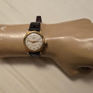 May include: A gold-tone wristwatch with a white face and black leather band. The watch face has black Roman numeral hour markers and black hands. The watch face also has the text "Junior" and "Swiss Made" printed on it.