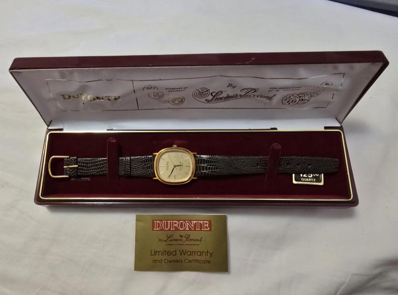 May include: A gold-tone watch with a black leather band in a burgundy box. The watch face is square and has gold-tone hands and markers. The box has a white interior and a label that reads "DuPonte by Swiss Perand Limited Warranty and Owners Certificate".