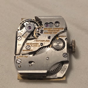 以下が含まれることがあります： 銀色の時計ムーブメントの裏側をクローズアップ。"Golden Watch Co. Precision - Swiss Made"と"Seventeen Jewels - Switzerland"の文字が見える。時計ムーブメントにはプッシュボタンと巻き上げ軸がある。
