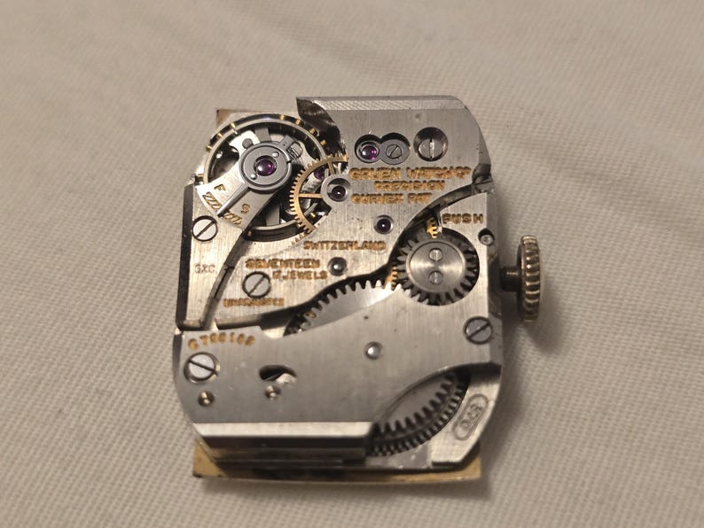 以下が含まれることがあります： 銀色の時計ムーブメントの裏側をクローズアップ。"Golden Watch Co. Precision - Swiss Made"と"Seventeen Jewels - Switzerland"の文字が見える。時計ムーブメントにはプッシュボタンと巻き上げ軸がある。