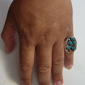 May include: A silver ring with an oval face, set with turquoise and coral stones. The ring is on a hand with tan skin, the fingers are visible, and the background is white.