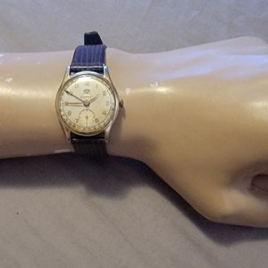 Może przedstawiać: Zegarek vintage z kopertą w kolorze srebrnym i kremową tarczą. Zegarek ma ciemnoniebieski pasek i jest prezentowany na dłoni manekina. Tarcza zegarka ma czarne cyfry i wskazówki.