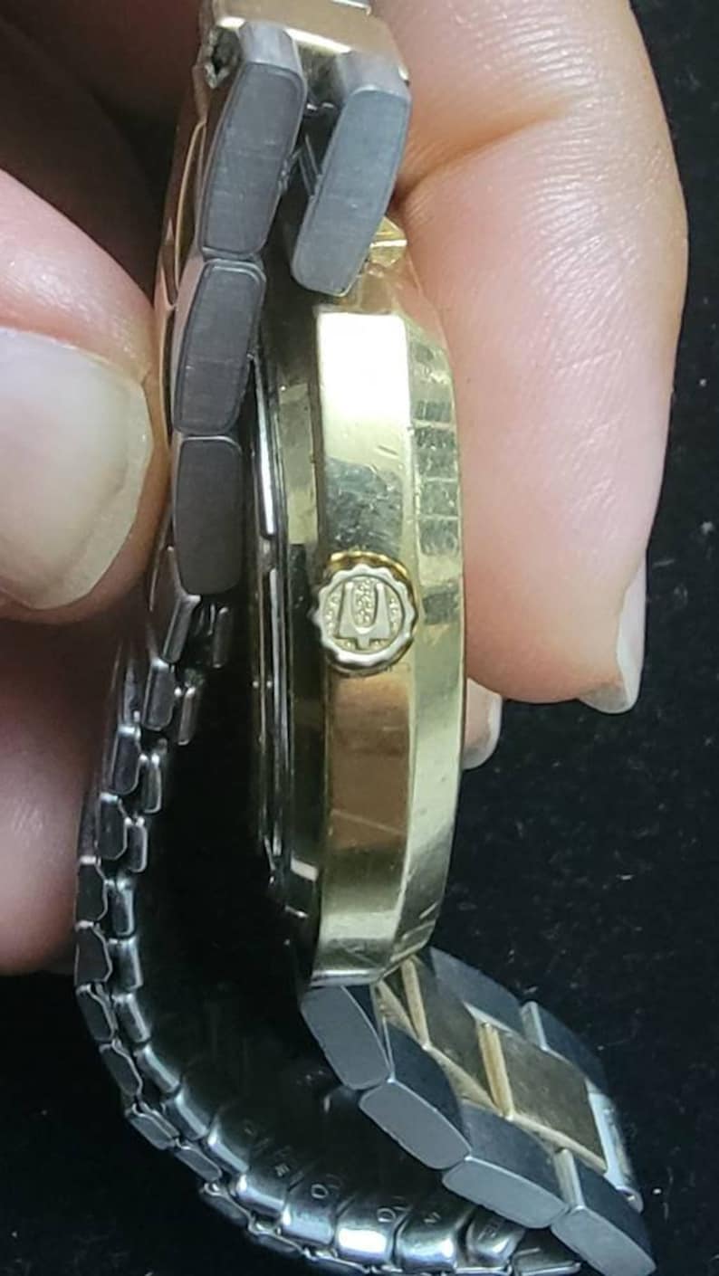 K&ouml;nnte beinhalten: Eine goldfarbene Armbanduhr mit einem silbernen Metallband. Das Zifferblatt der Uhr ist auf dem Bild nicht zu sehen. Die Uhr hat ein Logo an der Seite, auf dem "U" mit einer Krone dar&uuml;ber steht.