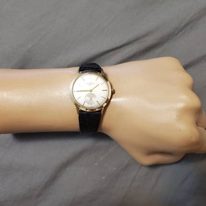May include: A gold wristwatch with a white face and black leather band. The watch has a simple design with black Roman numeral hour markers and a small seconds dial at the 6 o'clock position.