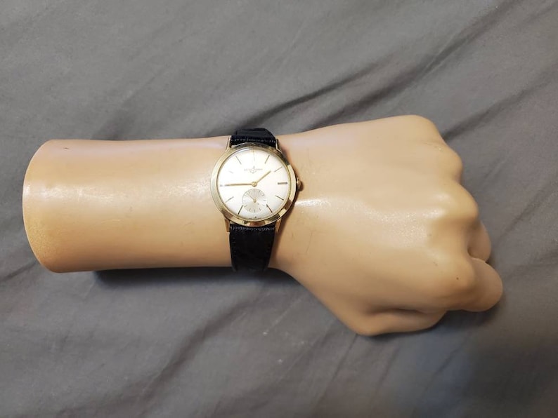 May include: A gold wristwatch with a white face and black leather band. The watch has a simple design with black Roman numeral hour markers and a small seconds dial at the 6 o'clock position.