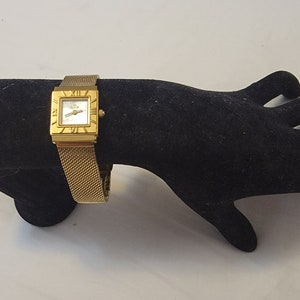 Könnte beinhalten: Eine goldfarbene Armbanduhr mit quadratischem Zifferblatt und einem geflochtenen Metallband. Das Zifferblatt hat römische Ziffern als Stundenmarkierungen und einen weißen Hintergrund.