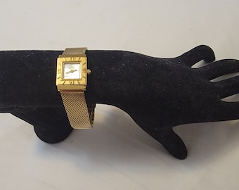 May include: A gold-tone wristwatch featuring a square case with Roman numeral markers on the bezel and a white dial. The watch has a flexible gold-tone mesh metal strap with a fine texture, providing a sleek and uniform appearance for the timepiece.