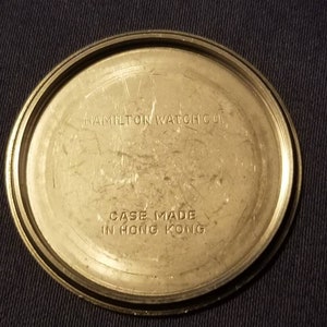 May include: A close-up of the back of a vintage Hamilton Watch Co. watch. The watch back is a circular, metallic color with the text "HAMILTON WATCH CO." and "CASE MADE IN HONG KONG" etched into the metal.