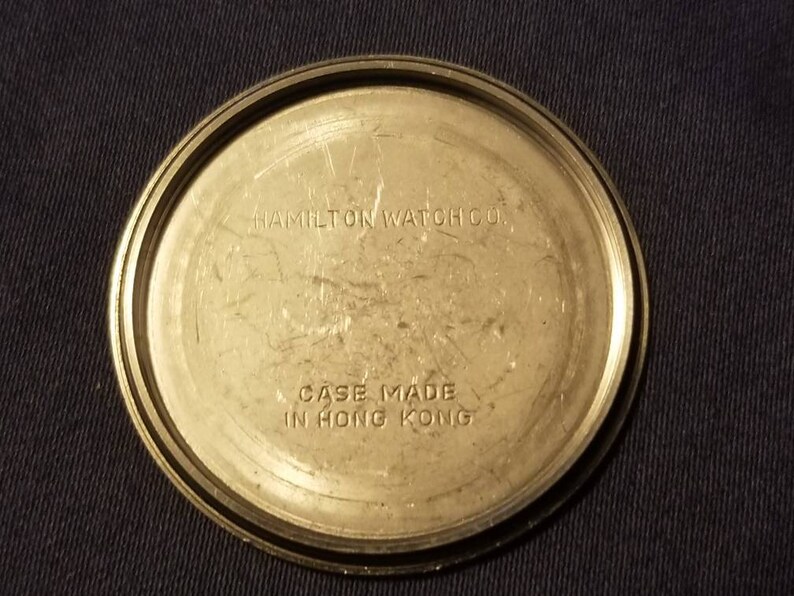 May include: A close-up of the back of a vintage Hamilton Watch Co. watch. The watch back is a circular, metallic color with the text "HAMILTON WATCH CO." and "CASE MADE IN HONG KONG" etched into the metal.