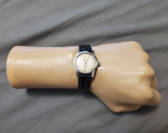 Può includere: Un orologio da polso argentato con quadrante bianco e cinturino in pelle nera. L'orologio ha una finestrella per la data e una lancetta dei secondi.
