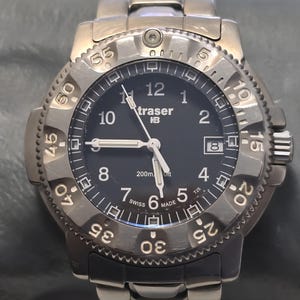 Puede incluir: Un reloj Traser P6500 Type 3 negro con un bisel plateado y marcas blancas. La esfera del reloj tiene un fondo negro con n&uacute;meros y manecillas blancas. El reloj es resistente al agua hasta 200 metros.