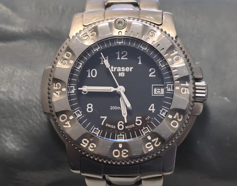 Puede incluir: Un reloj Traser P6500 Type 3 negro con un bisel plateado y marcas blancas. La esfera del reloj tiene un fondo negro con n&uacute;meros y manecillas blancas. El reloj es resistente al agua hasta 200 metros.