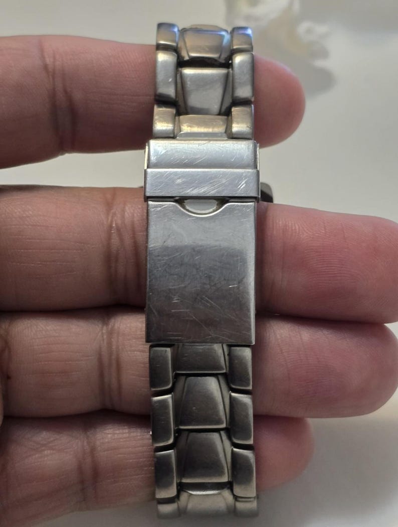 Puede incluir: Una correa de reloj de metal plateado con un cierre. La correa est&aacute; hecha de piezas de metal enlazadas.