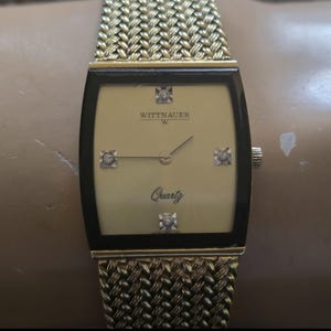 May include: A gold-toned Wittnauer watch with a square face and a gold metal band. The watch face has a gold background with black Roman numeral hour markers and a quartz movement. The watch has four small diamonds on the face.