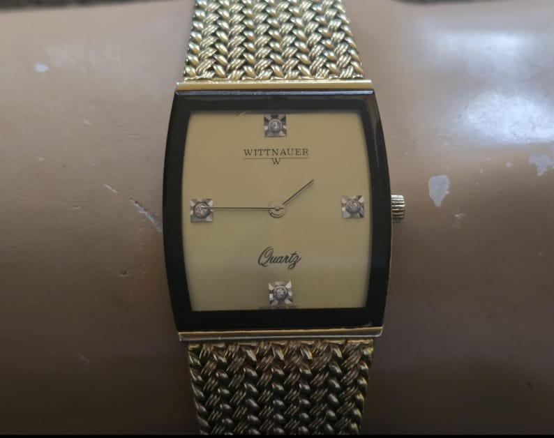 May include: A gold-toned Wittnauer watch with a square face and a gold metal band. The watch face has a gold background with black Roman numeral hour markers and a quartz movement. The watch has four small diamonds on the face.