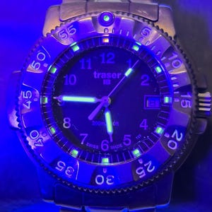 Puede incluir: Un reloj Traser 100m negro con una esfera negra y agujas e &iacute;ndices luminiscentes verdes. El reloj tiene un bisel negro con n&uacute;meros blancos y una correa de acero inoxidable.