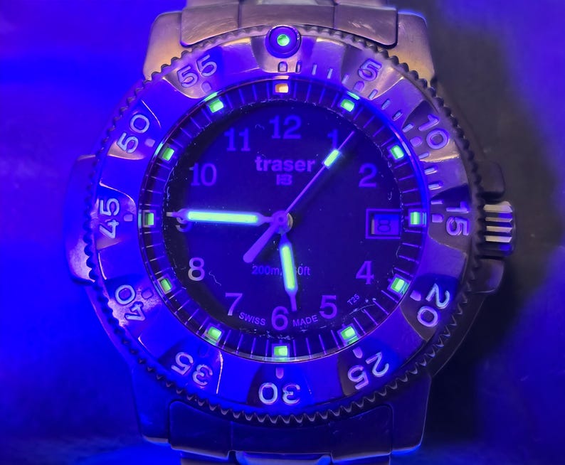 Puede incluir: Un reloj Traser 100m negro con una esfera negra y agujas e &iacute;ndices luminiscentes verdes. El reloj tiene un bisel negro con n&uacute;meros blancos y una correa de acero inoxidable.