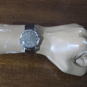 May include: A wristwatch with a round silver-tone case and a dark brown textured leather strap. The watch face is grey with silver-tone hands, silver-tone hour markers, and the numbers 2, 4, 6, 8, 10, and 12 displayed in a simple font.