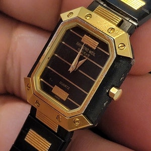 May include: A gold and black wristwatch with a square face. The watch face has a black background with gold accents and the text "RAYMOND WEIL GENEVE" and "QUARTZ".