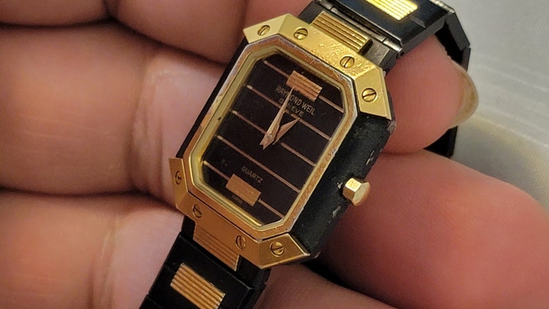 May include: A gold and black wristwatch with a square face. The watch face has a black background with gold accents and the text "RAYMOND WEIL GENEVE" and "QUARTZ".