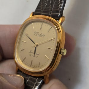 May include: A gold-tone watch with a square face and a brown leather band. The watch face has a white background with black hands and black Roman numeral hour markers. The watch face also has the text "Duforte Swiss Quartz" printed on it.