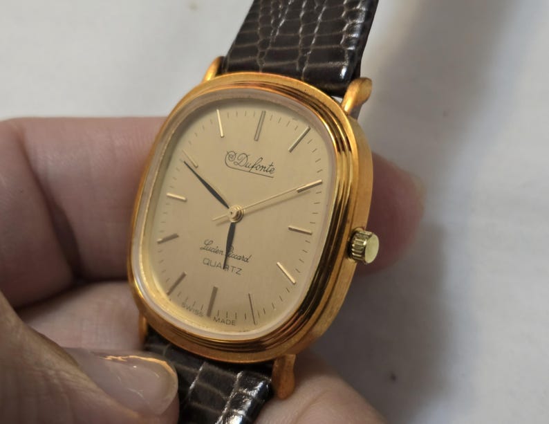 May include: A gold-tone watch with a square face and a brown leather band. The watch face has a white background with black hands and black Roman numeral hour markers. The watch face also has the text "Duforte Swiss Quartz" printed on it.