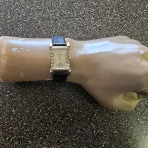 Puede incluir: Un reloj dorado con esfera blanca y correa de cuero negra. El reloj está en la muñeca de un maniquí.