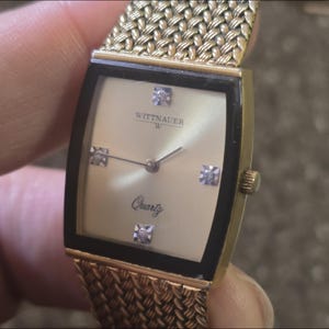 May include: A gold-toned Wittnauer W quartz watch with a black face and diamond accents. The watch has a gold mesh band.