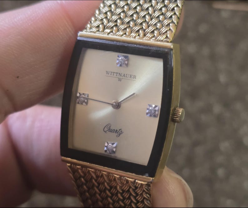 May include: A gold-toned Wittnauer W quartz watch with a black face and diamond accents. The watch has a gold mesh band.