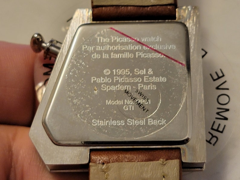 May include: The back of a silver watch with a brown leather band. The watch face is engraved with the text "The Picasso watch Par autorisation exclusive de la famille Picasso. &copy; 1995, Sel & Pablo Picasso Estate Spadem - Paris SWISS MOVEMENT Model No: PS1 GTI Stainless Steel Back".
