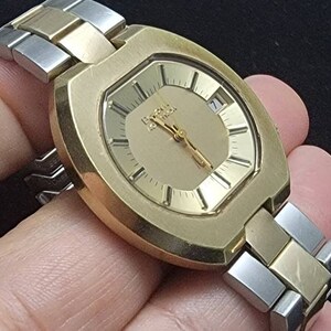 K&ouml;nnte beinhalten: Eine goldfarbene Armbanduhr mit quadratischem Zifferblatt und einem zweifarbigen Armband aus Silber und Gold. Das Zifferblatt hat einen goldenen Hintergrund mit schwarzen Markierungen und einem Datumsfenster.