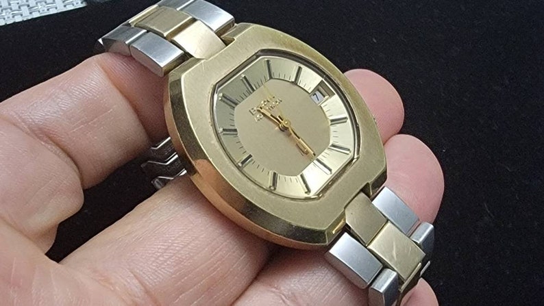 K&ouml;nnte beinhalten: Eine goldfarbene Armbanduhr mit quadratischem Zifferblatt und einem zweifarbigen Armband aus Silber und Gold. Das Zifferblatt hat einen goldenen Hintergrund mit schwarzen Markierungen und einem Datumsfenster.