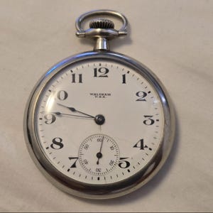 Può includere: Un orologio da tasca d'argento con un quadrante bianco e indici orari in numeri romani neri. L'orologio ha una lancetta dei secondi e un piccolo quadrante secondario per i secondi. Il quadrante dell'orologio ha anche il testo "WALTHAM U.S.A."
