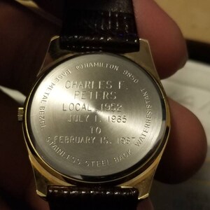 May include: A gold-toned wristwatch with a brown leather band. The watch back is engraved with text including "CHARLES F. PETERS" and dates from 1952 to 1987. The watch has a stainless steel back and a base metal bezel.