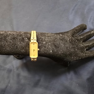 Könnte beinhalten: Eine goldfarbene Armbanduhr mit rechteckigem Zifferblatt und römischen Ziffern. Die Uhr wird auf einer dunkelgrauen handförmigen Skulptur präsentiert. Das Armband ist strukturiert. Der Hintergrund ist ein dunkelblauer Stoff.