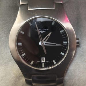 May include: A stainless steel wristwatch with a black dial. The watch face has silver hands and markers, with the brand name "LONGINES" at the top. The date is displayed at the 6 o'clock position. The watch has a silver strap.