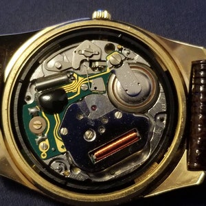 May include: Close-up of a gold-toned wristwatch with a brown leather band. The watch's inner workings are visible, showcasing intricate mechanical components, including a circuit board, battery, and various gears. The watch face is open, revealing the detailed internal mechanisms.