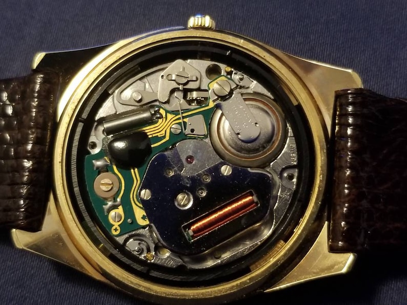 May include: Close-up of a gold-toned wristwatch with a brown leather band. The watch's inner workings are visible, showcasing intricate mechanical components, including a circuit board, battery, and various gears. The watch face is open, revealing the detailed internal mechanisms.
