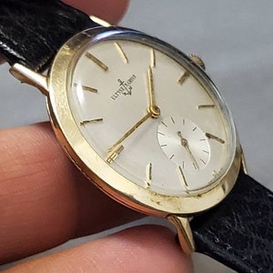May include: A gold-tone wristwatch with a white face and black leather band. The watch face has black Roman numeral hour markers and a small seconds dial at the 6 o'clock position. The watch face also has the text "ULYSSE NARDIN" printed on it.