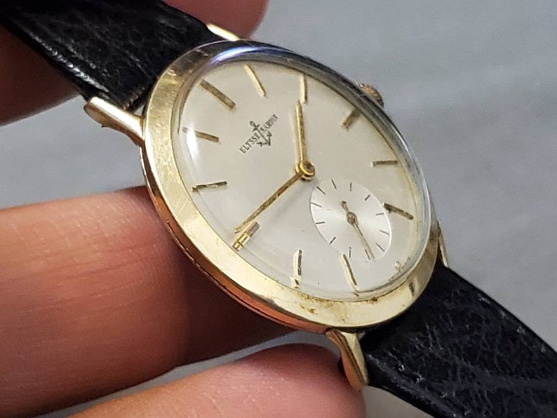 May include: A gold-tone wristwatch with a white face and black leather band. The watch face has black Roman numeral hour markers and a small seconds dial at the 6 o'clock position. The watch face also has the text "ULYSSE NARDIN" printed on it.