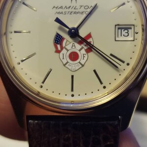 May include: A gold-toned Hamilton Masterpiece watch with a cream-colored face. The watch features the date 13, black hands, and a firefighter emblem. The watch has a brown leather band.