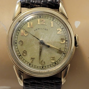 May include: A gold-toned wristwatch with a white face and black hands. The watch has black Roman numeral hour markers and a black second hand. The watch face also has the word "Hamilton" printed on it.